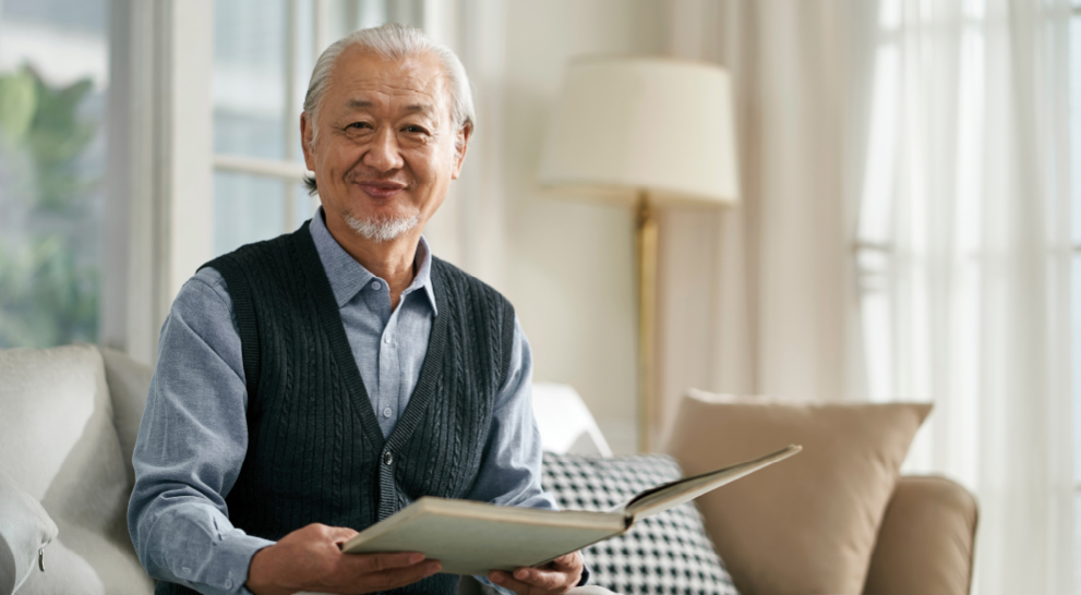 A senior holds a book in his hands and smiles from sitting on a couch at an affordable senior living community.