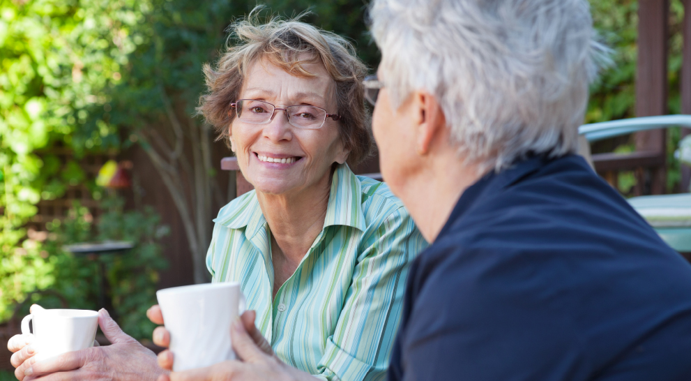 Two senior women sit on a bench with cups of coffee in hand. They smile at one another. One of the women is out of focus.