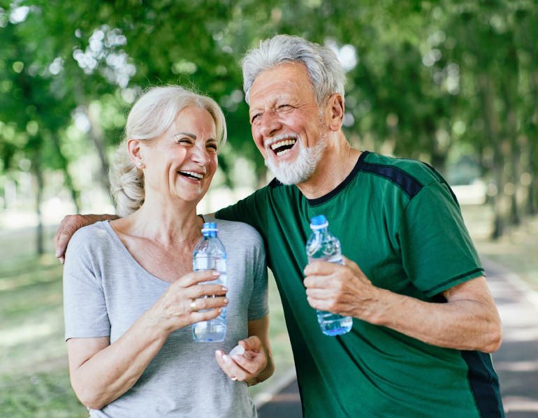 A senior couple with water bottles laugh together while outside in the sun.